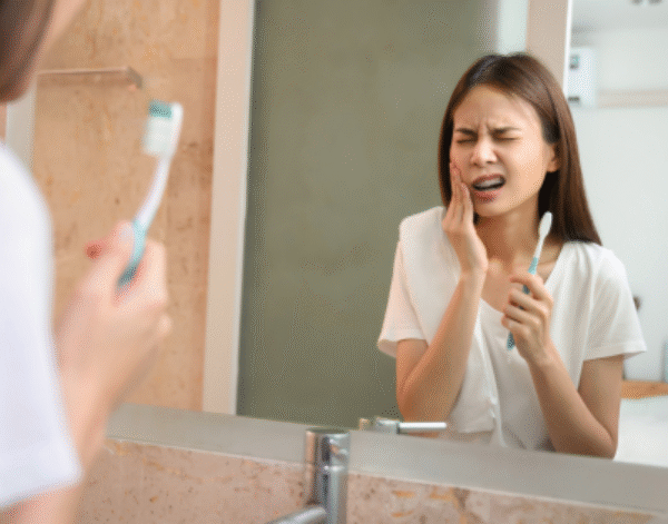 Woman brushing teeth in mirror, showing pain from sensitive teeth