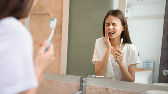 Woman brushing teeth in mirror, showing pain from sensitive teeth